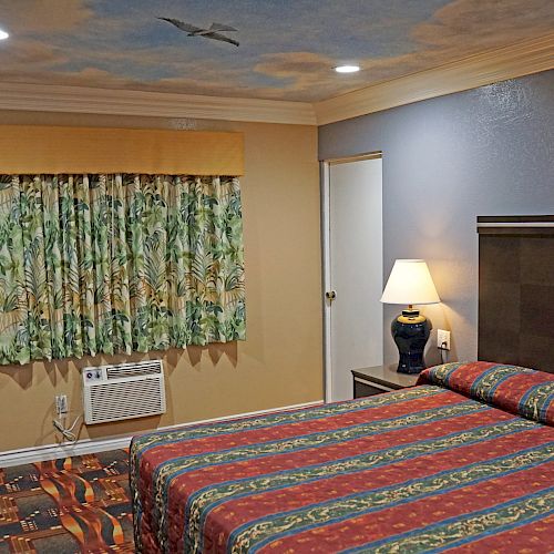 A hotel room with a bed covered in a colorful striped blanket, a lamp, floral curtains, a door, and air conditioner, ceiling with a bird design.