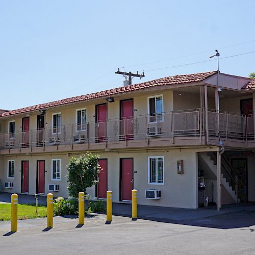 The image shows a two-story motel with red doors, a staircase, and a parking lot. Yellow bollards and greenery are in the foreground.