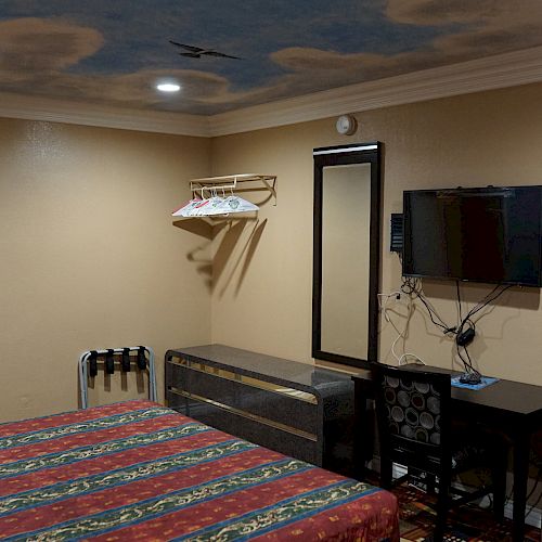 A room with a bed, a wall-mounted TV, a desk and chair, a mini fridge, and a wall painting. There’s a rack with hangers and a mirror.