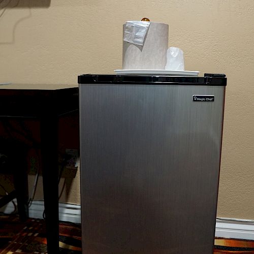 A small refrigerator is placed against the wall, with paper towels and a cup on top. A table and painting are also partially visible in the image.