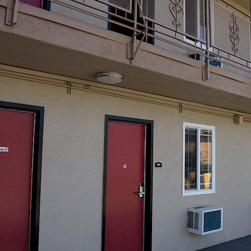 The image shows the exterior of a motel or apartment building with two floors, red doors, and air conditioning units mounted on the lower walls.