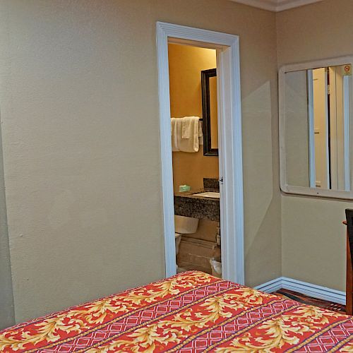 The image shows a hotel room with a bed, a wall-mounted TV, a desk with a chair, an open door to a bathroom, and a wall mirror.