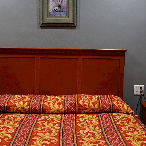 The image shows a bed with a patterned red and yellow bedspread, a wooden headboard, a nightstand with a lamp, and a framed picture on the wall.