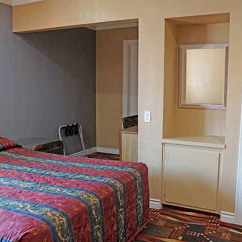 A hotel room with a multi-colored bedspread, a wall with hangers, a small counter space, and a bathroom entrance with towels visible.