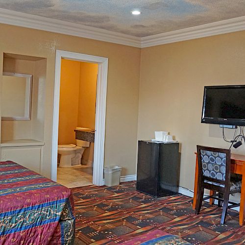 The image depicts a hotel room with a bed, a TV mounted on the wall, a desk with a chair, and an open door to a bathroom in the background.