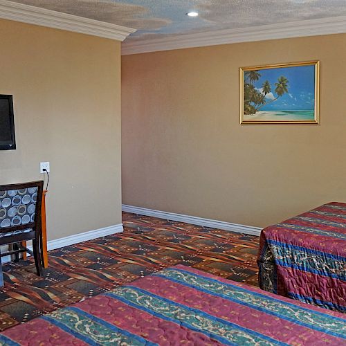 The image shows a hotel room with two beds, a wall-mounted TV above a desk and chair, a framed beach painting, and patterned carpet flooring.