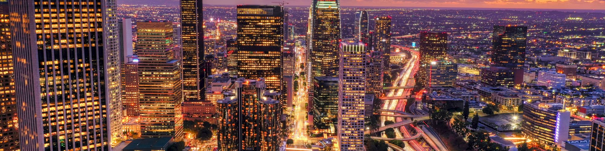 The image depicts a vibrant cityscape at dusk, featuring illuminated skyscrapers, busy roads with light trails, and a dramatic sky with clouds.