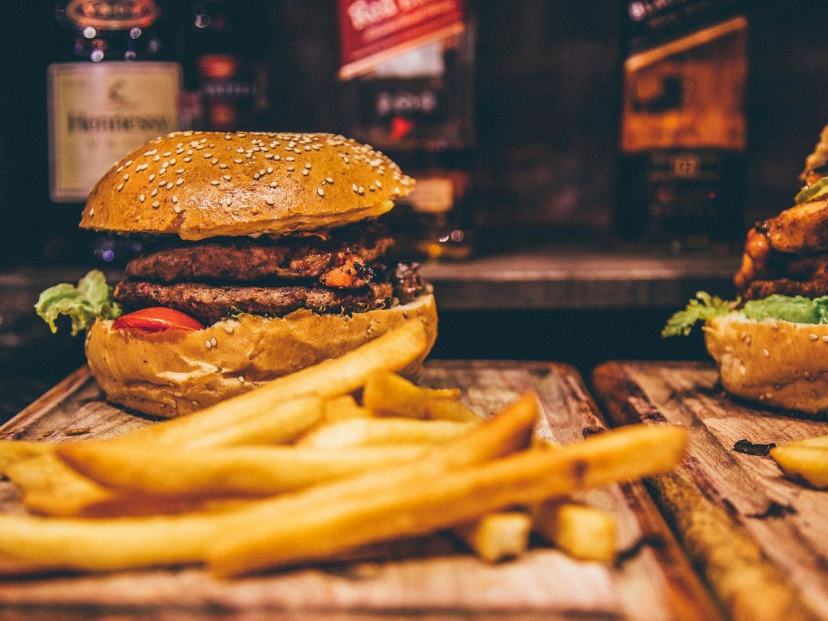 A burger with sesame seed bun and fries on a wooden board is in focus, with blurred bottles in the background.