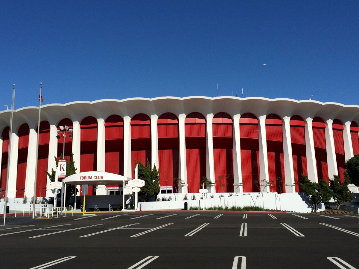 The image shows a large, circular, red and white arena with numerous pillars and an adjacent empty parking lot under a clear blue sky.