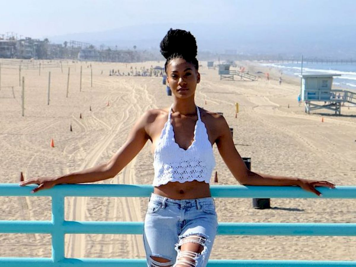 A person stands on a pier railing, with a sandy beach and ocean view behind them, wearing a white top and ripped jeans.