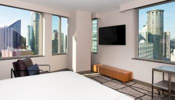 This image shows a modern hotel room with large windows offering a city view, a mounted TV, a chair, a bed, and a table.