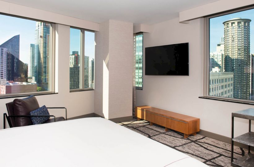 This image shows a modern hotel room with large windows offering a city view, a mounted TV, a chair, a bed, and a table.