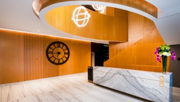 A modern lobby or reception area with a marble desk, a large wall clock, wooden paneling, and decorative lighting fixtures and flowers.