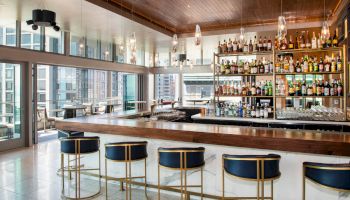 A modern, well-lit bar with a large selection of liquor bottles, high ceilings, blue and gold barstools, and a view of the city through large windows.