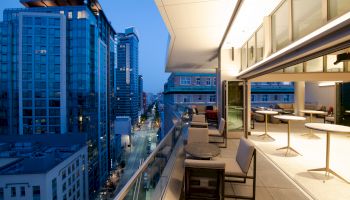 A modern urban balcony with tables and chairs overlooks a cityscape filled with tall buildings, captured during dusk with city lights.
