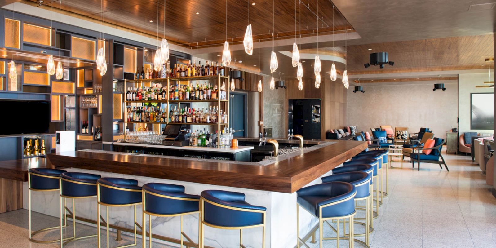 A modern bar with a wooden countertop, blue cushioned bar stools, a full liquor shelf, pendant lights, and a spacious seating area in the background.