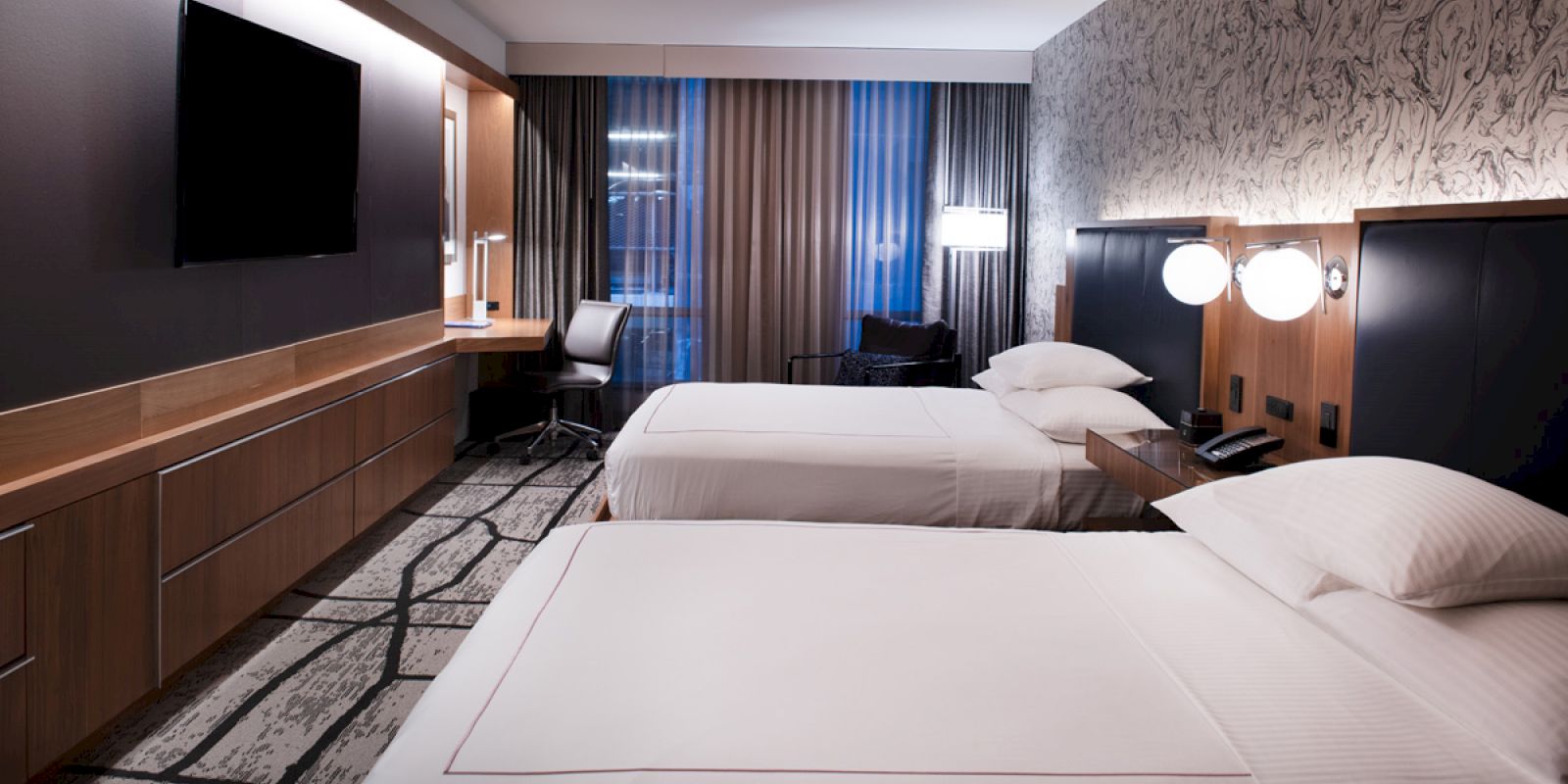 This image shows a modern hotel room with two beds, a desk and chair, a large TV, and stylish decor and lighting.