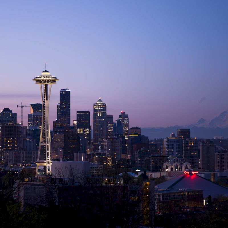 A city skyline at dusk with prominent buildings including the Space Needle, and a mountain in the background under a clear sky.