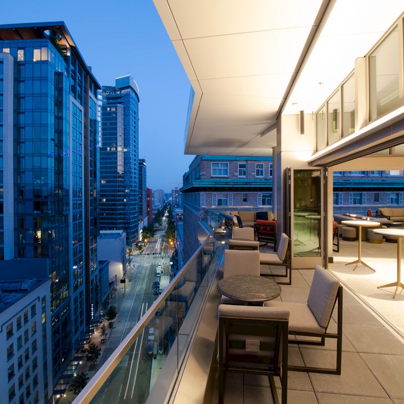 A modern high-rise balcony with seating and tables overlooks a city street lined with other tall buildings at dusk or early evening.