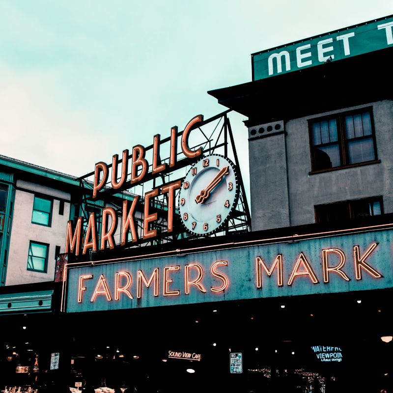 A public market with neon signs reads "PUBLIC MARKET" and "FARMERS MARKET." A clock is visible in the center. A nearby building says "MEET THE."