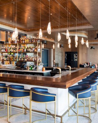 A modern bar with a central counter, blue bar stools, hanging lights, wide windows, and a stocked bar with shelves of various bottles.