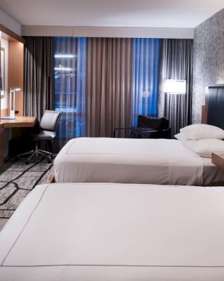 The image shows a modern hotel room with two beds, a TV, a desk with a chair, and large windows with curtains, in a contemporary design.