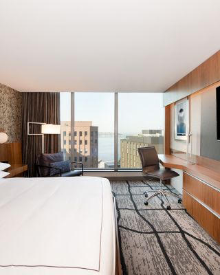 A modern hotel room featuring a large bed, a wall-mounted TV, a desk with a chair, a window with a city view, and contemporary decor.
