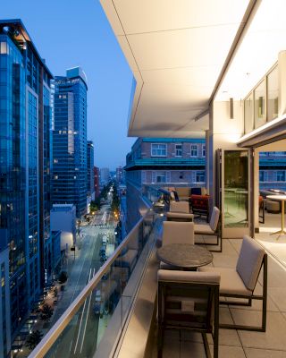 A modern balcony overlooks a cityscape with tall buildings; it has seating areas and tables near large windows with a nighttime view.