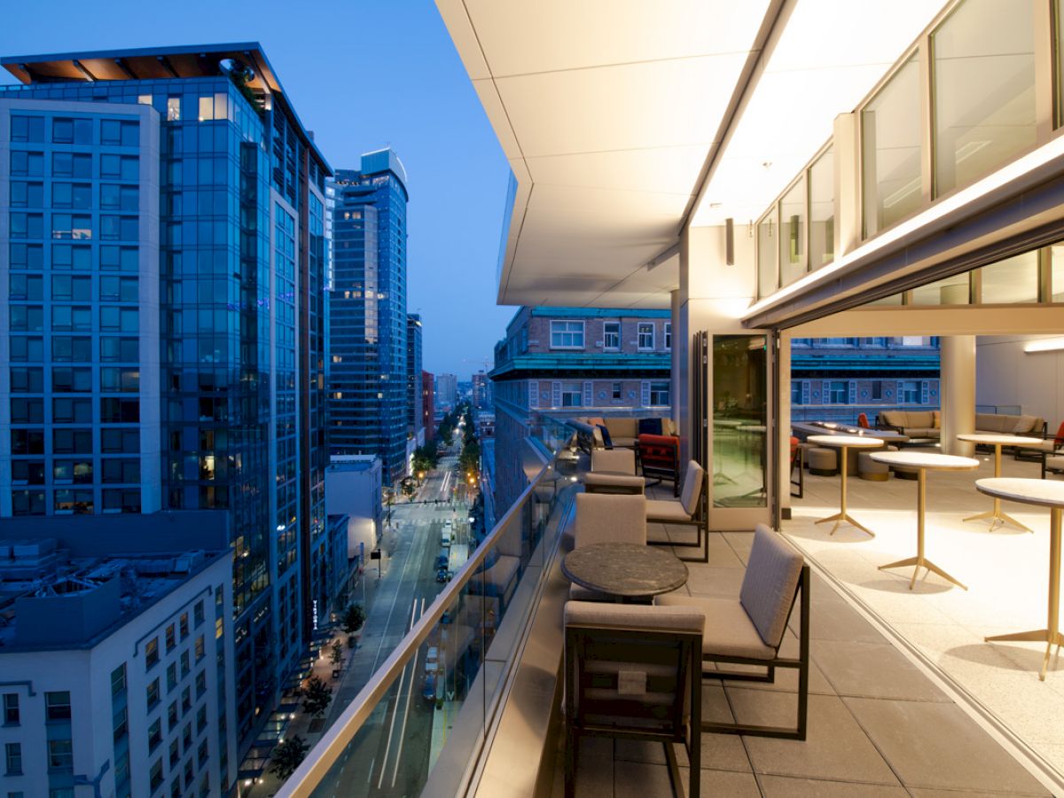 A rooftop patio with modern furniture overlooks a cityscape with tall buildings and a lit street at dusk, featuring large windows and tables.
