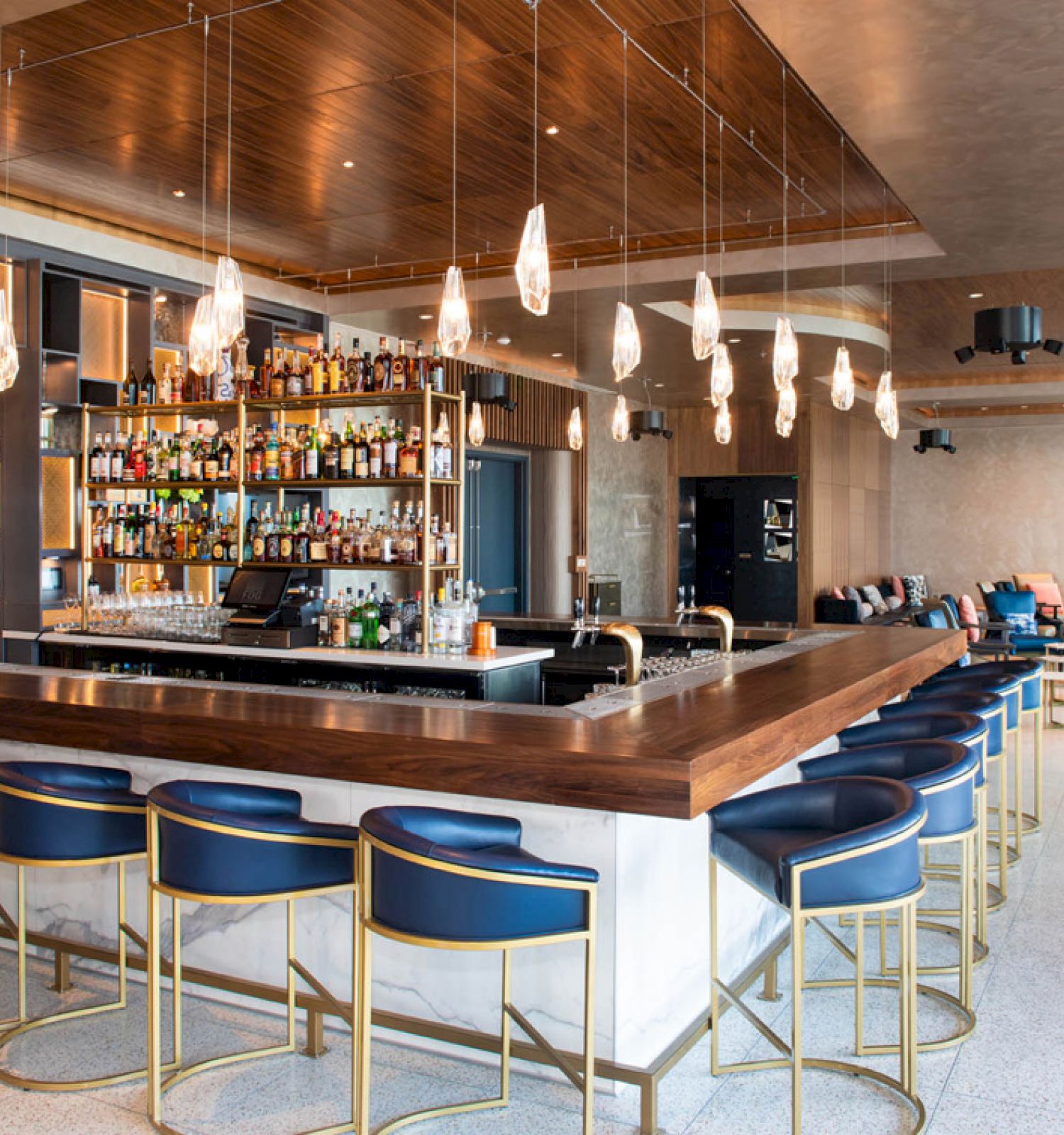 A modern bar featuring a wooden counter with blue barstools, elegant lighting, and an extensive selection of bottles behind the bar.