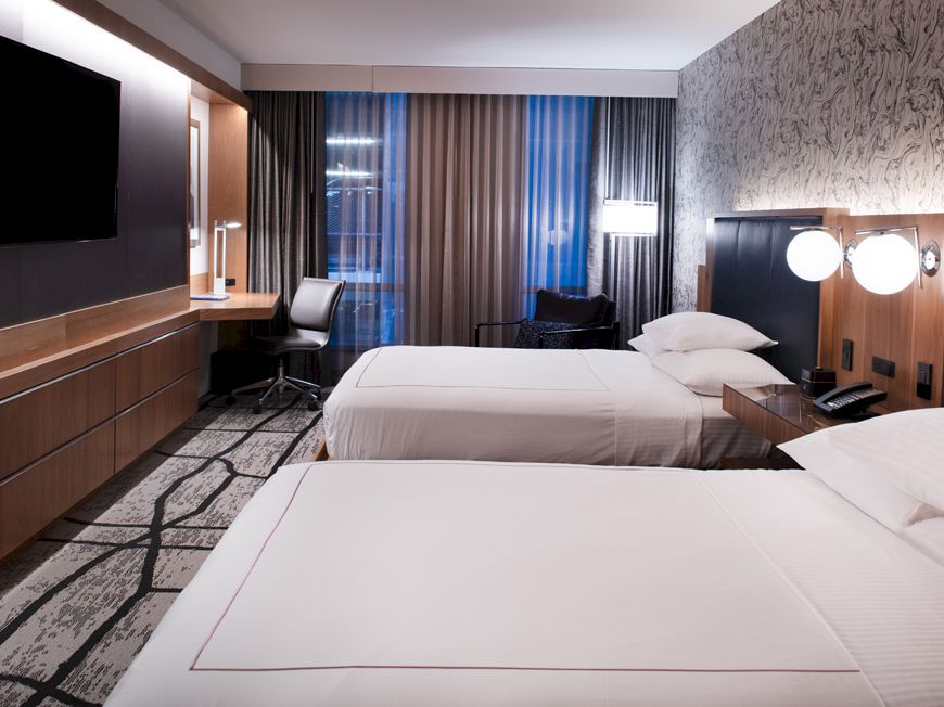 The image shows a modern hotel room with two beds, a flat-screen TV, a desk area with a chair, lamps, and large window curtains.