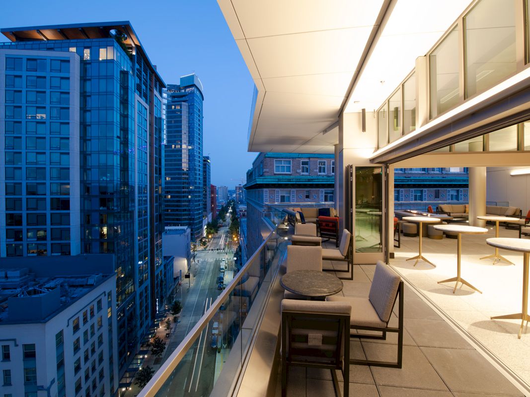 A modern cityscape at dusk with tall buildings, seen from a balcony with seating and tables, offering a panoramic city view.