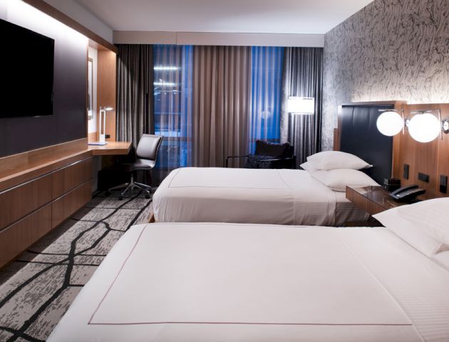 The image shows a modern hotel room with two double beds, a wall-mounted TV, a desk, and stylish lighting and decor, creating a cozy ambiance.