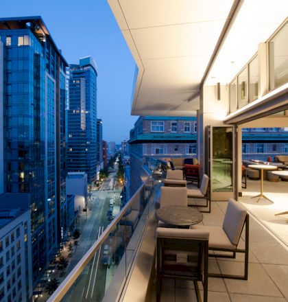 This image shows a modern cityscape with tall buildings, viewed from a high balcony with tables and chairs, at evening twilight.