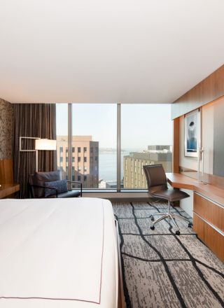 This is a modern hotel room featuring a large bed, a desk with a chair, a flat-screen TV, and a view of buildings outside the window.
