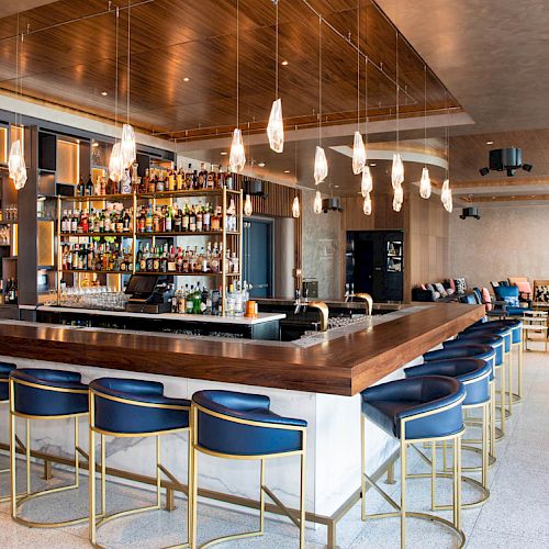 A modern bar with a large counter, blue barstools, an array of drinks on shelves, and stylish lighting hanging from the ceiling.