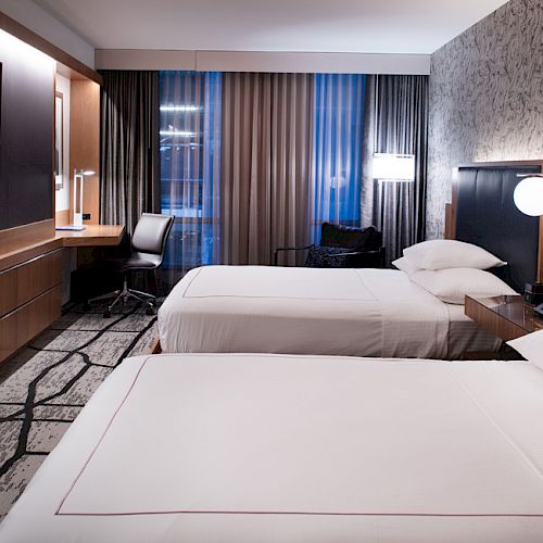 A modern hotel room with two neatly made beds, a flat-screen TV, a desk, and contemporary decor featuring a patterned accent wall and ample lighting.