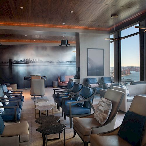 The image shows a modern lounge area with large windows offering a city and waterfront view, and comfortable seating arranged around small tables.