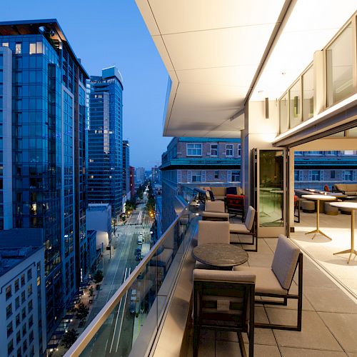 A modern cityscape at dusk featuring a high-rise building with a furnished balcony overlooking a street and more tall buildings.