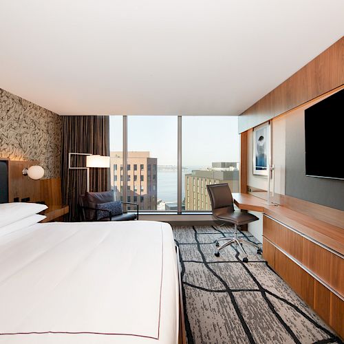 A modern hotel room with a king-size bed, large TV, desk, chair, and a view of city buildings through the window ends the sentence.