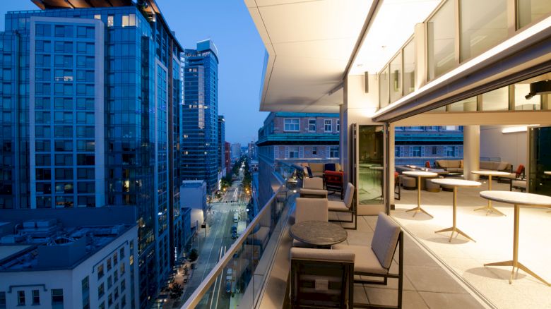 A modern cityscape at dusk, viewed from a high balcony with seating and tables. High-rise buildings and lit windows are visible in the background.
