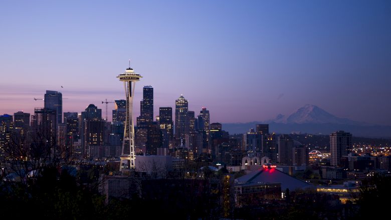 A skyline featuring the Space Needle in Seattle at dusk with city buildings and Mount Rainier visible in the background.