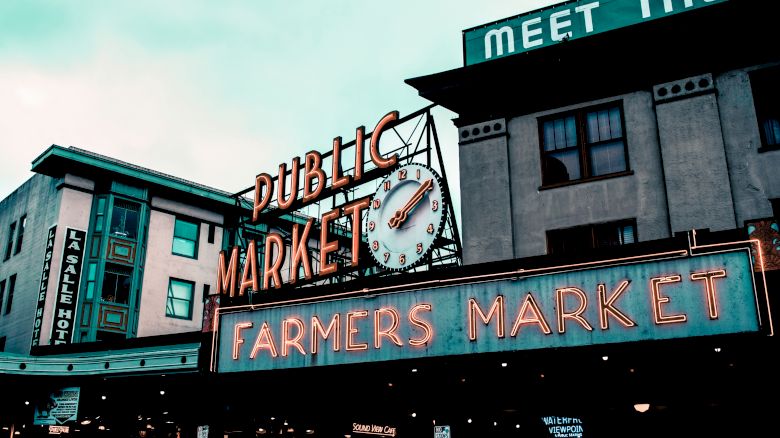 The image features a neon sign for a "Public Market" and "Farmers Market" against the backdrop of urban buildings, with a large clock.