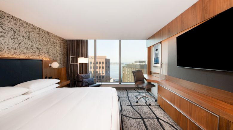 This image shows a modern hotel room with a large bed, a desk, a chair, a flat-screen TV, and a view of a cityscape through the window.