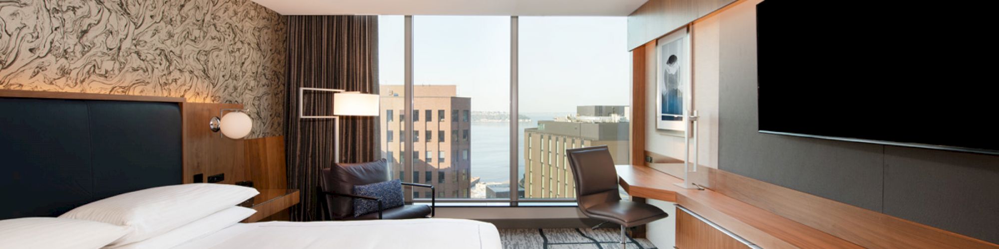 A modern hotel room with a large bed, wall-mounted TV, desk, and a chair by the window overlooking a cityscape and waterfront view.