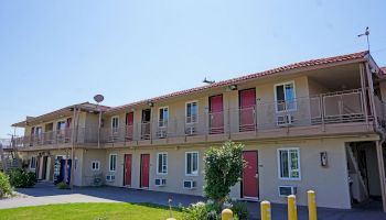 The image shows a two-story motel with red doors, beige walls, and outdoor staircases. There is greenery in the foreground and a vending machine.