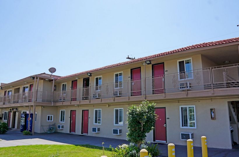 The image shows a two-story motel with red doors, beige walls, and outdoor staircases. There is greenery in the foreground and a vending machine.