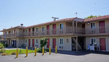 The image shows a two-story motel with red doors, beige walls, exterior staircases, and multiple windows, situated in a clear weather setting.