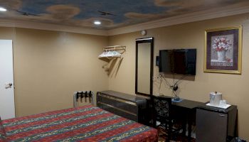 A hotel room with a bed, wall-mounted TV, mini-fridge, painting, and a coat rack. Ceiling has blue cloud patterns and lighting. Ends the sentence.