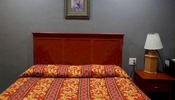 The image shows a bed with a red and yellow patterned bedspread, a wooden headboard, a bedside table with a lamp, and a framed picture on the wall.